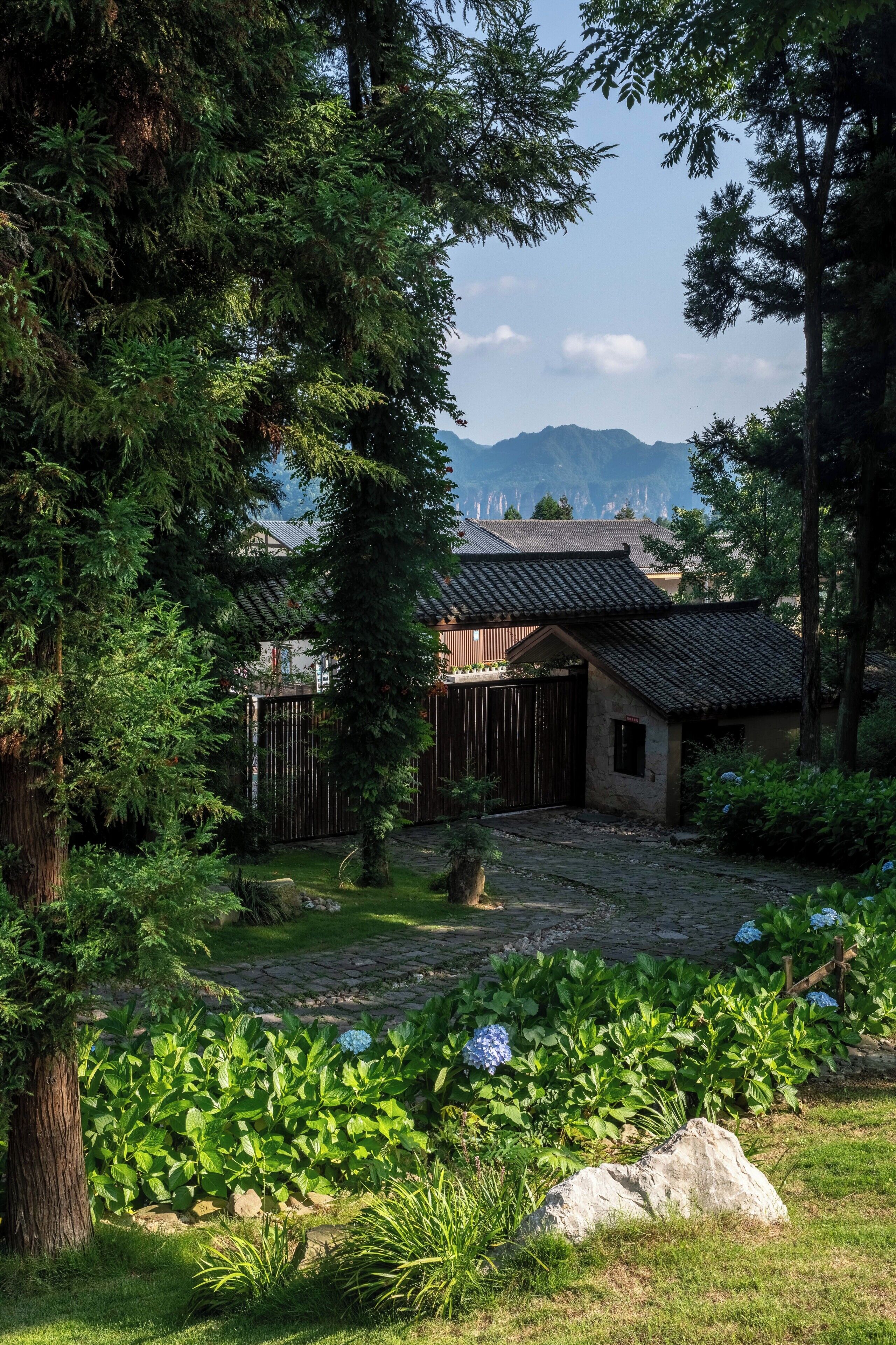 Un bâtiment de style japonais traditionnel, avec un toit recouvert de tuiles, entouré d’une végétation luxuriante et bordé d’un sentier en pierre.