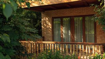 A wooden deck with a railing, a brick wall, and a window with a view of greenery.