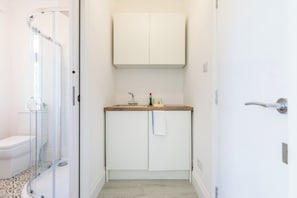 A narrow hallway with a glass-enclosed shower, a white toilet, and a small kitchenette with a sink and cabinet. - Light Filled First Floor Studio with Bay Window (Greater London)