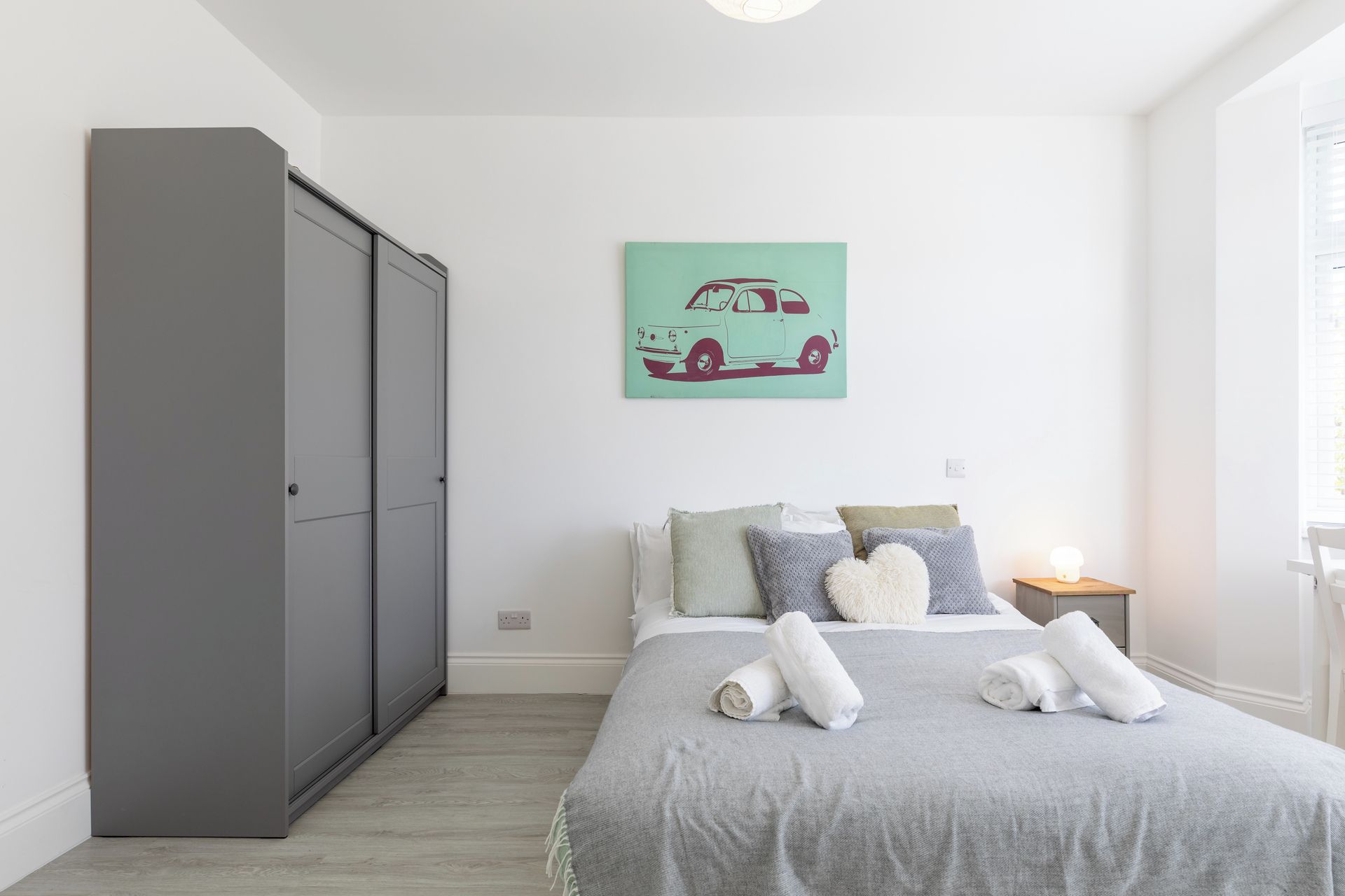 A bedroom with a grey wardrobe, a bed with grey bedding, a teal car illustration on the wall, and a bedside lamp.