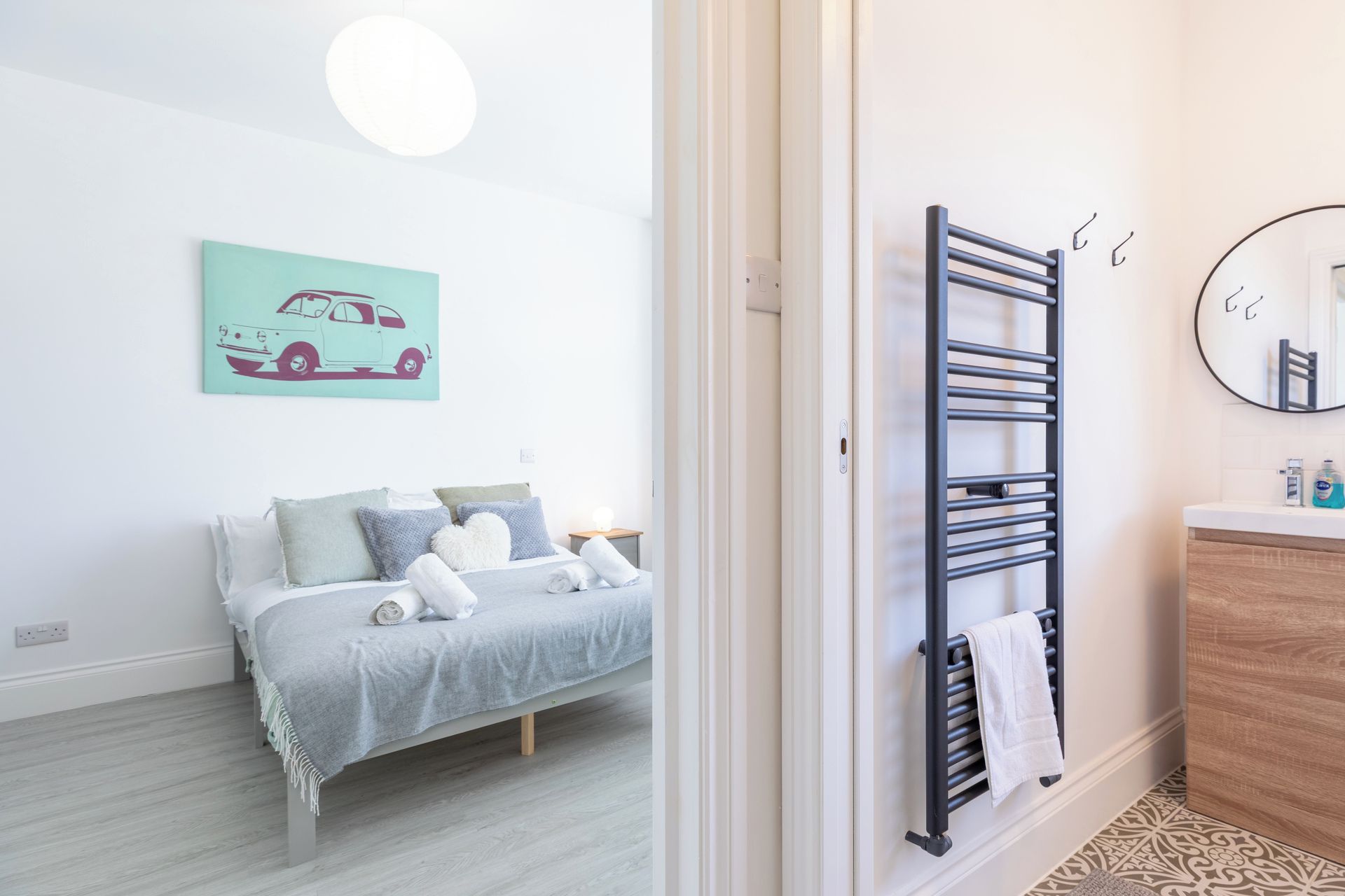 A bedroom with a bed, a bedside table, a wall-mounted radiator, a bathroom with a mirror, and a wall art of a car.