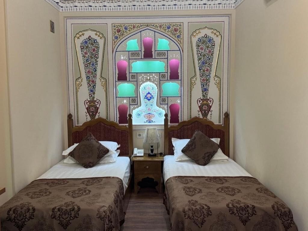 Habitación de hotel con dos camas, una mesita de noche y una pared decorativa con diseños complejos.