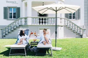 Two lounge chairs with a table and umbrella in front of a building with a staircase and a sign reading 'Verena's Guesthouse'.
