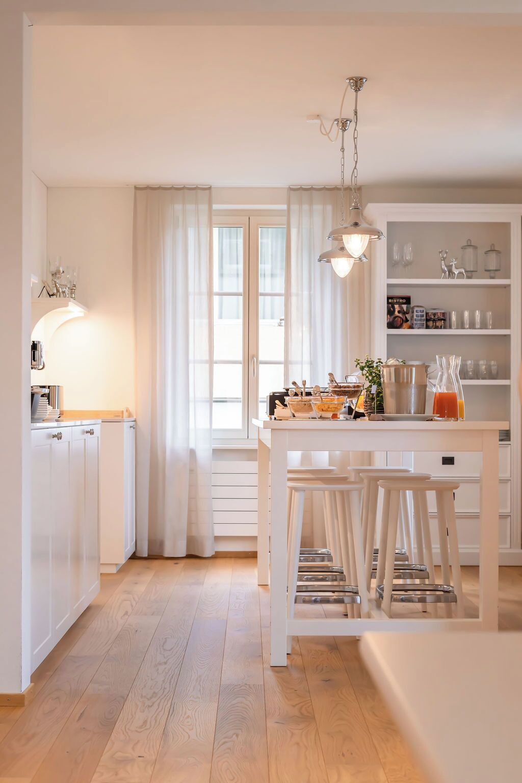 Une cuisine moderne avec des meubles blancs, un coin repas doté de tabourets et une grande fenêtre agrémentée de rideaux.