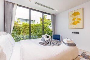 A hotel room with a large bed, a blue chair, a wall-mounted artwork, and a view of greenery outside. - Botanica Forestique 4br Luxury Retreat (Choeng Thale)