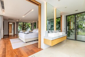 A modern bedroom with a large bed, a bathroom with a large mirror and a bathtub, and a view of greenery outside. - Botanica Forestique 4br Luxury Retreat (Choeng Thale)