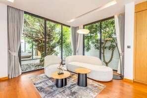 Een moderne woonkamer met een bank, twee bijzettafels, een vloerkleed en grote ramen met gordijnen.