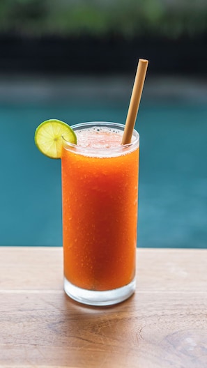 A tall glass of orange beverage with a straw and a lime slice, placed on a wooden surface with a blurred pool in the background. - Lavana Ubud (Ubud)