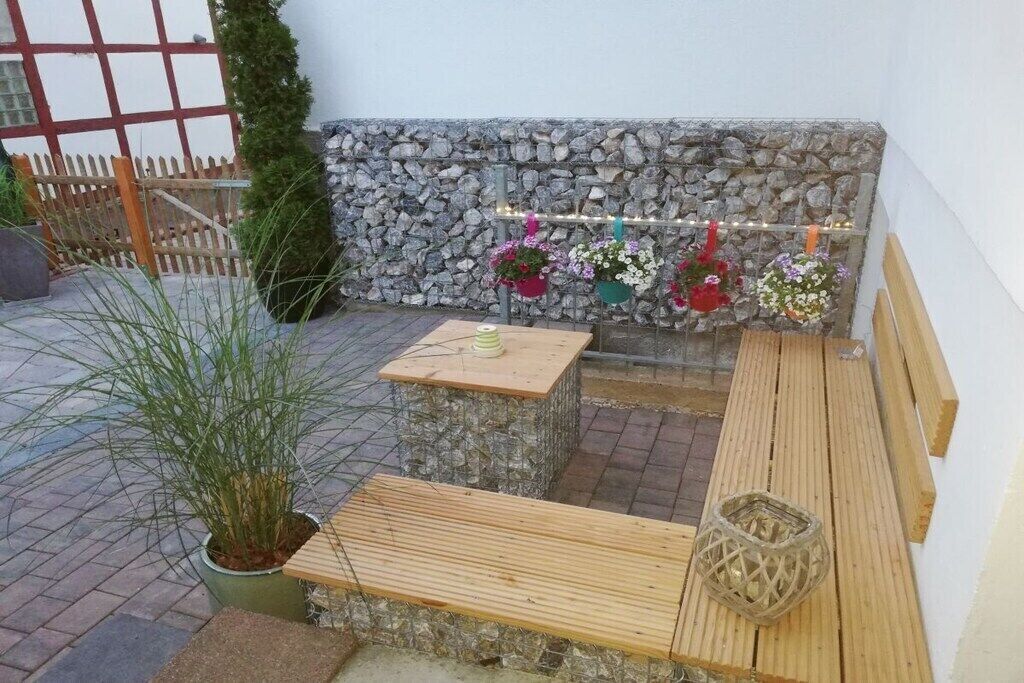 Eine Steinsäule mit hängenden Blumenkörben, einer Holzbank und einem kleinen Tisch mit einer Kerze.