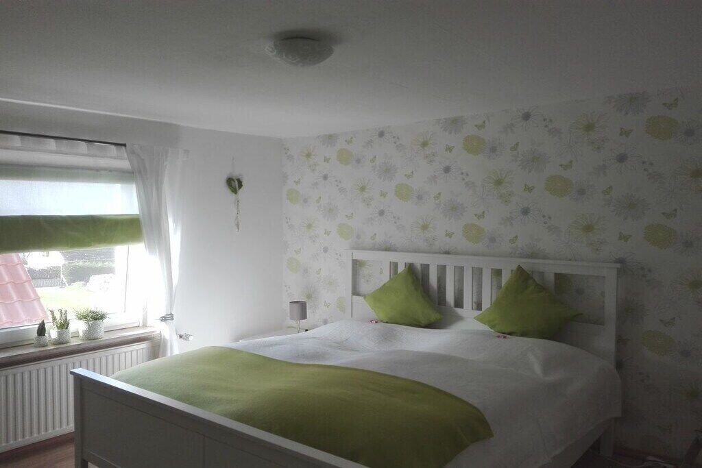 Une chambre à coucher avec un lit blanc, des coussins verts, une fenêtre avec des rideaux et un mur orné d’un motif floral.