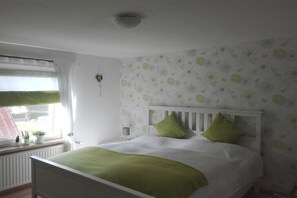 Une chambre à coucher avec un lit blanc, des coussins verts, une fenêtre avec des rideaux et un mur orné d’un motif floral.