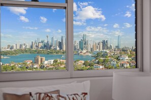 A room with a large window offering a view of a city skyline, a body of water, and a hill with greenery. - Penthouse Near Taronga Zoo w/ Iconic Harbour Views (Mosman)