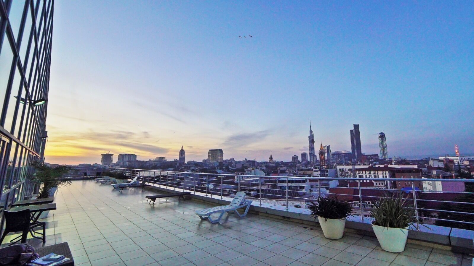 Een terras op het dak met uitzicht op de skyline van de stad, lounge stoelen en plantenbakken.