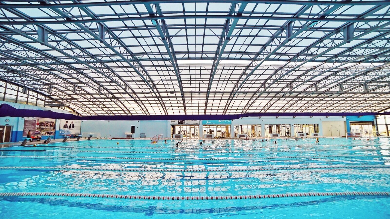 An indoor swimming pool with multiple lanes, a high ceiling with a grid-like structure, and various poolside amenities.