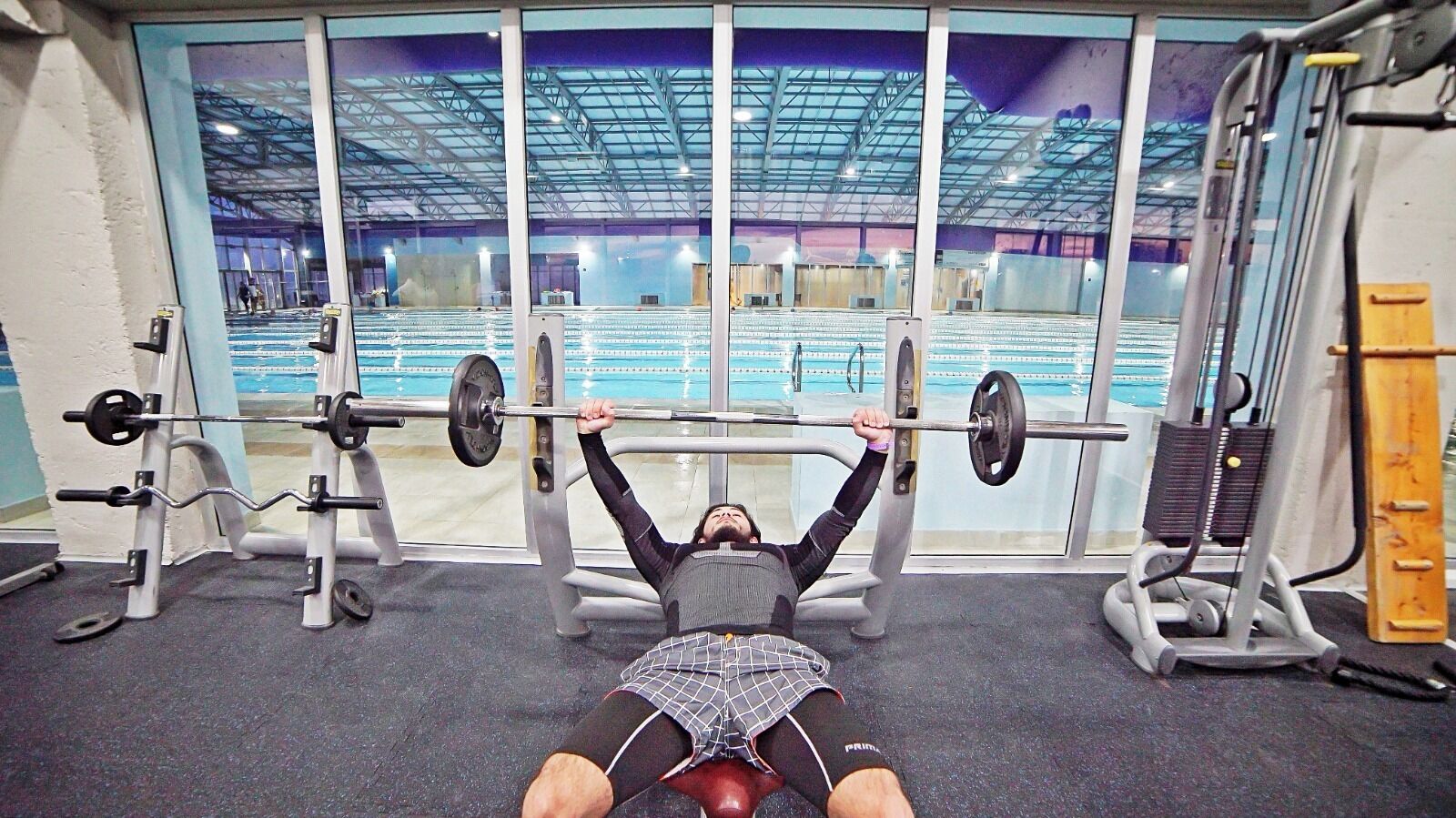 Un gimnasio con una estación de press de banca, un soporte para pesas y un ventanal con vistas a una zona de piscina interior.