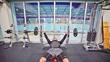 Un gimnasio con una estación de press de banca, un soporte para pesas y un ventanal con vistas a una zona de piscina interior.