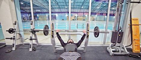 A gym with a bench press setup, a weight rack, and a large window overlooking an indoor pool area.