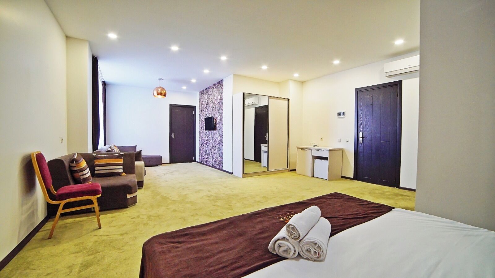 A hotel room with a bed, a seating area with a chair and cushions, a small desk, and a mirror.