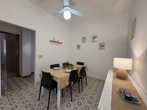 Un coin repas avec une table dressée pour quatre personnes, des chaises, un ventilateur de plafond et une étagère murale agrémentée d’objets décoratifs.