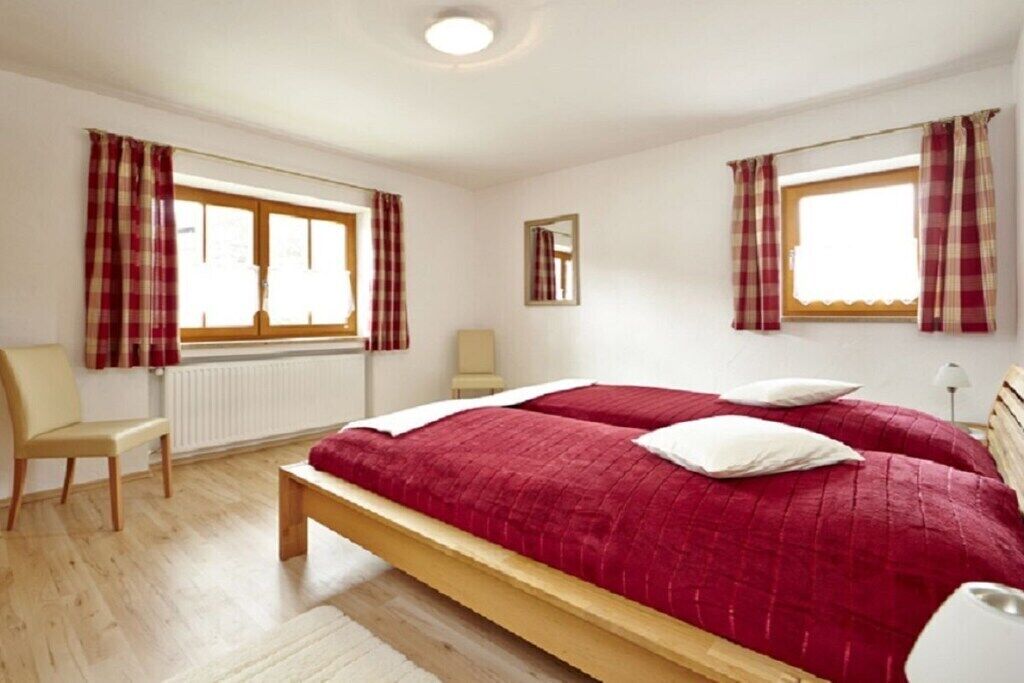 Quarto com duas camas, uma cadeira, mesa de cabeceira e janela com cortinas.