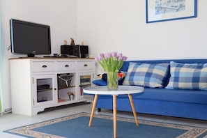 Ein Wohnzimmer mit einem blauen Sofa, einer weißen Kommode, einem Flachbildfernseher und einer Vase mit Blumen auf einem kleinen runden Tisch.