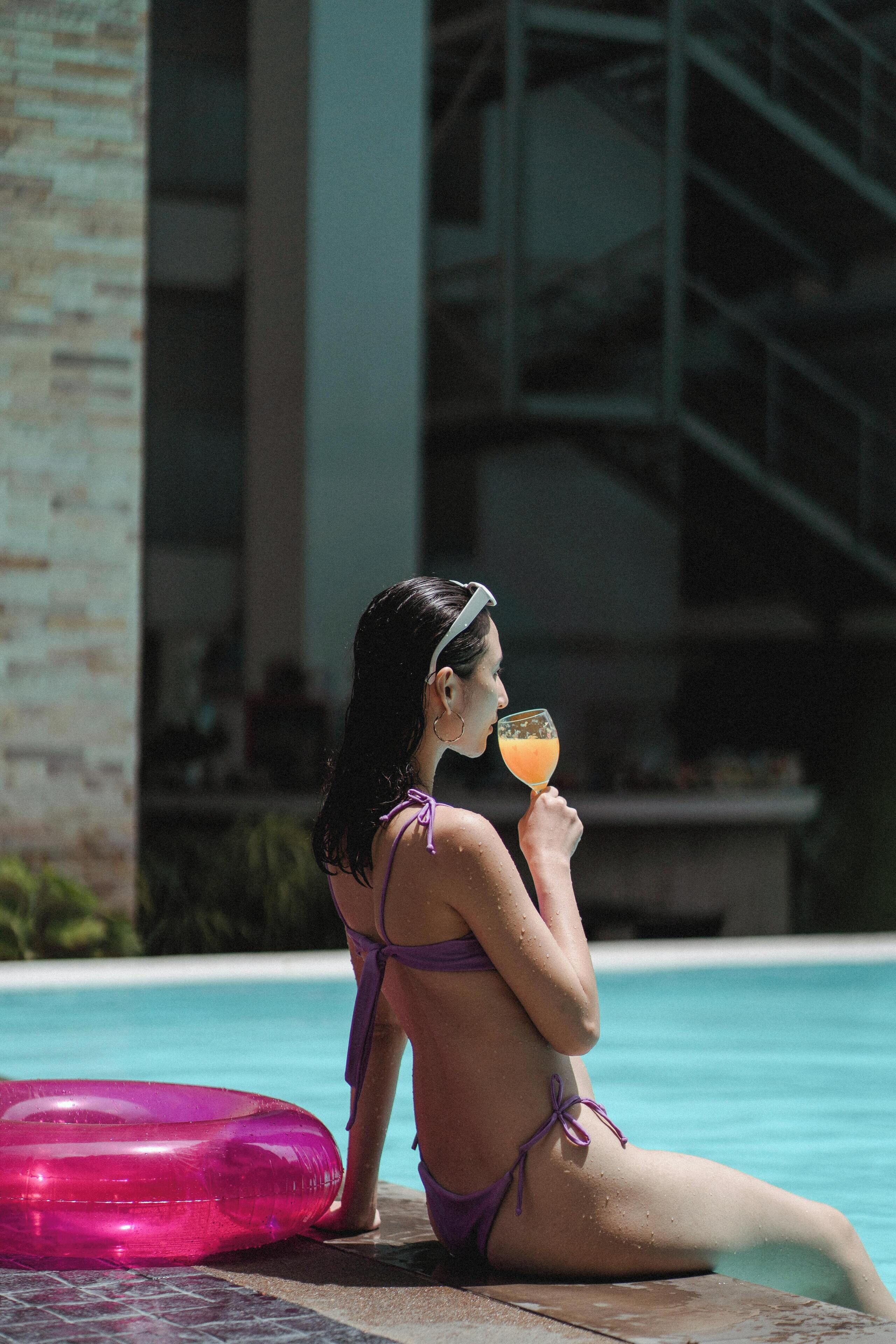 Une personne vêtue d’un bikini violet, assise au bord d’une piscine, tenant un verre à la main, avec un flotteur gonflable rose à proximité et un bâtiment en arrière-plan.