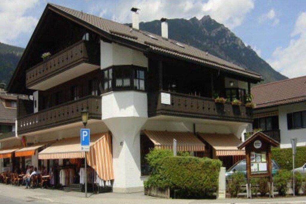 A traditional chalet-style building with a balcony, a visible parking sign, and a mountain backdrop.