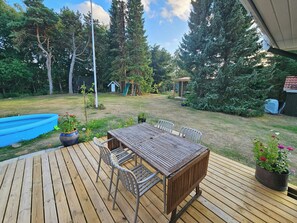 Eine Holzterrasse mit Tisch und Stühlen, umgeben von einer grünen Rasenfläche mit Bäumen und einem blauen Swimmingpool.