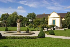Ein gepflegter Garten mit einer Fontäne, geschnittenen Hecken und einem kleinen Gebäude mit grüner Tür.