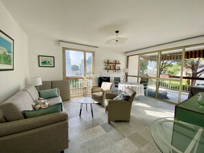 A spacious living room with a sofa, armchairs, a small table, and a glass coffee table. There are large windows providing a view of the outdoors. - 2-room apartment in a luxury residence—with a pool (Mandelieu-la-Napoule)
