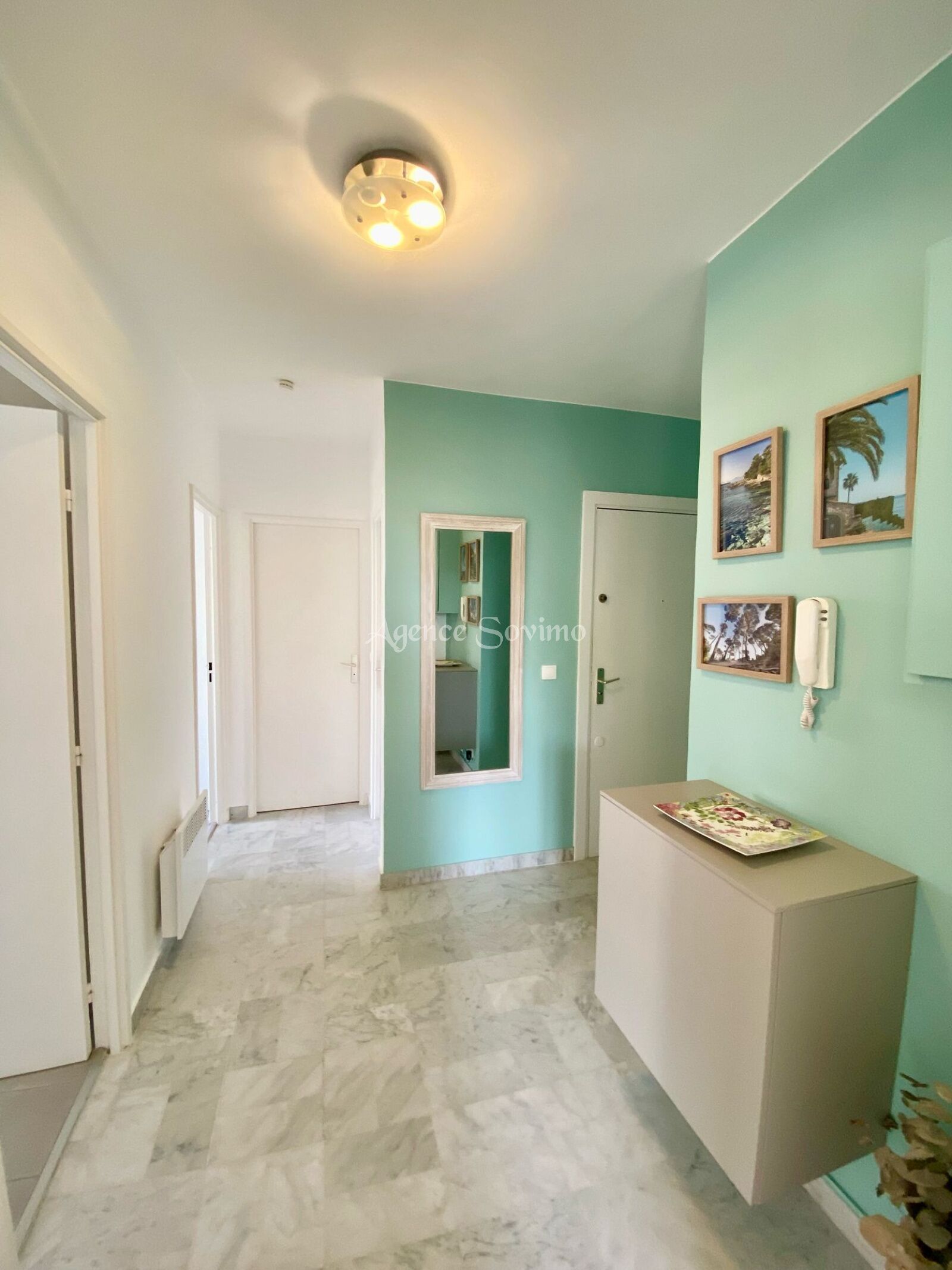 A hallway with a marble floor, a door at the end, and a wall with framed pictures and a telephone.