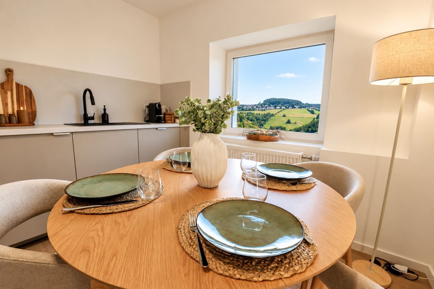 Ein Esstisch ist mit Tellern, Gläsern und einer Vase mit Grünpflanzen gedeckt, mit Blick auf eine Landschaft durch ein Fenster.