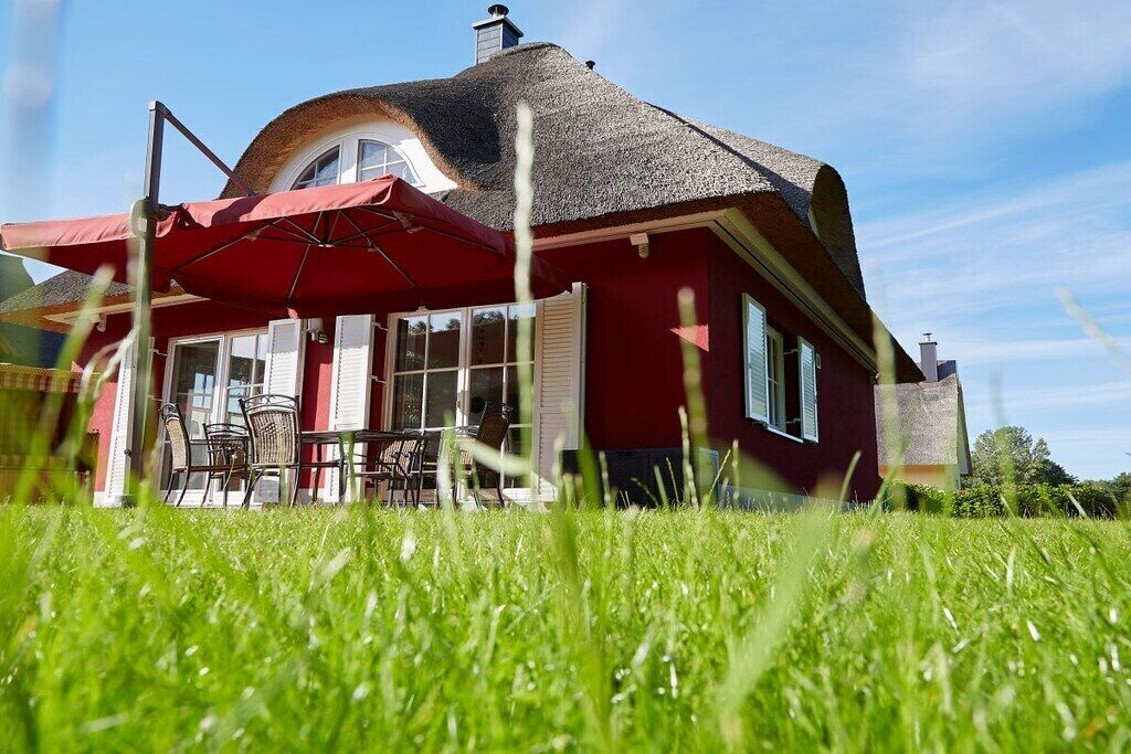 Une maison rouge avec un toit de chaume, un coin repas avec des meubles d’extérieur et un ciel bleu azur.