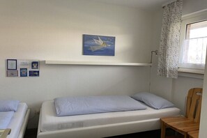 A single-bed room with a white headboard, a wooden chair, a window with a patterned curtain, and a wall-mounted shelf with a framed picture. - 4 in the Kiebitzwiese house (Borkum)
