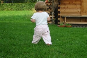 Un enfant se tient debout sur l’herbe, dans un jardin, avec une structure en bois en arrière-plan.
