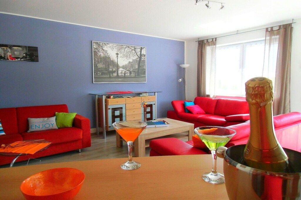 Een woonkamer met rode banken, een bijzettafeltje en drankjes. Aan de muur hangt een schilderij en er is een raam met gordijnen.