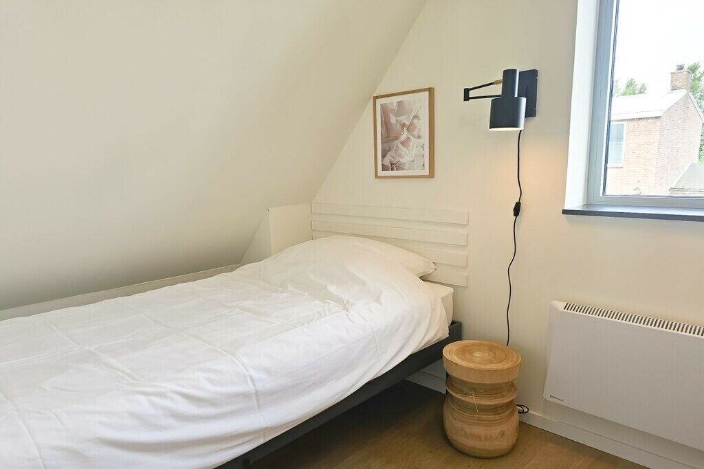 Uma cama de solteiro com colchão e cabeceira brancos, uma mesa de cabeceira de madeira, um aplique fixado na parede e uma janela com vista para edifícios.
