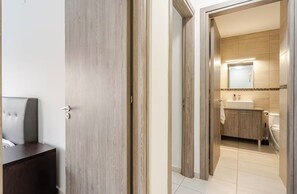 Ein Hotelzimmer mit Holztür, einem Bett mit Kopfteil und Blick ins Badezimmer mit Waschbecken und Spiegel.