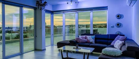 A modern living room with a black sofa, a coffee table, and a view of the city through large glass doors.