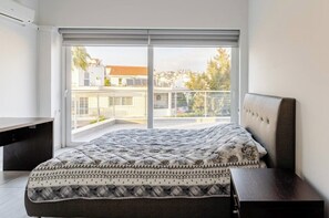 A bedroom with a bed, a bedside table, a window with a view of buildings, and a ceiling fan.