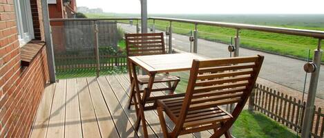 A wooden deck with a table and chairs, overlooking a road and green landscape.