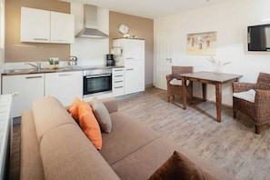 A compact living space with a kitchenette, a sofa, a dining table with chairs, and a television mounted on the wall. - Holiday apartment in Juist with swimming pool (Juist)