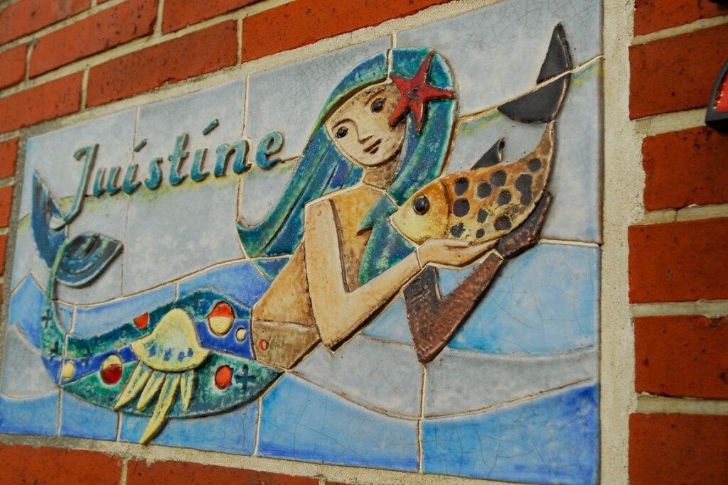 Ein mit Fliesen ausgelegtes Wandbild mit einer Meerjungfrau und Fischen, das den Schriftzug 'Justine' zeigt.