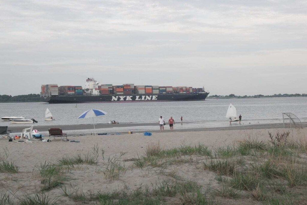 Een vrachtschip met 'NYK LINE' op de zijkant, afgemeerd aan een strand waar mensen lopen en strandstoelen en parasols staan.
