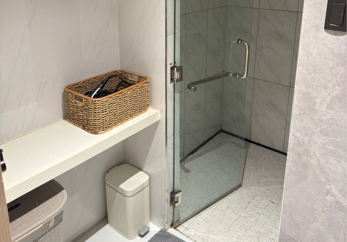 Un baño con mampara de vidrio, una cesta de mimbre en un estante y un cubo de basura.