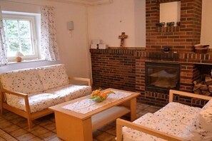 A cozy living room with a brick fireplace, a sofa, a coffee table with fruit, and a window with curtains. - Guesthouse with 4 bedrooms (Berge OT Börstel)