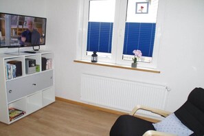 Ein Zimmer mit einem Fernseher auf einem weißen Schrank, einer weißen Heizung, einem Fenster mit blauen Jalousien, einem schwarzen Stuhl und einem kleinen Regal mit Büchern und einer Pflanze.