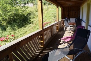 Um deck de madeira com móveis para área externa, incluindo cadeiras e uma mesa, com vista para um jardim com folhagem e flores.