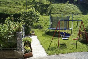 Ein Spielplatz mit Trampolin und Schaukeln, umgeben von einer Steinmauer und Grünflächen.