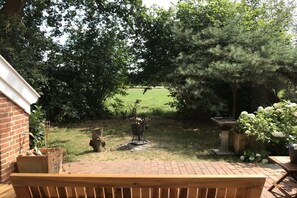 A patio with a brick floor, a wooden bench, and a view of a grassy yard with trees and a birdbath.